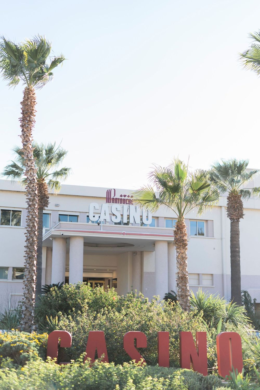 The white casino building facade features tall palm trees and red 'CASINO' lettering in the landscaped entrance.