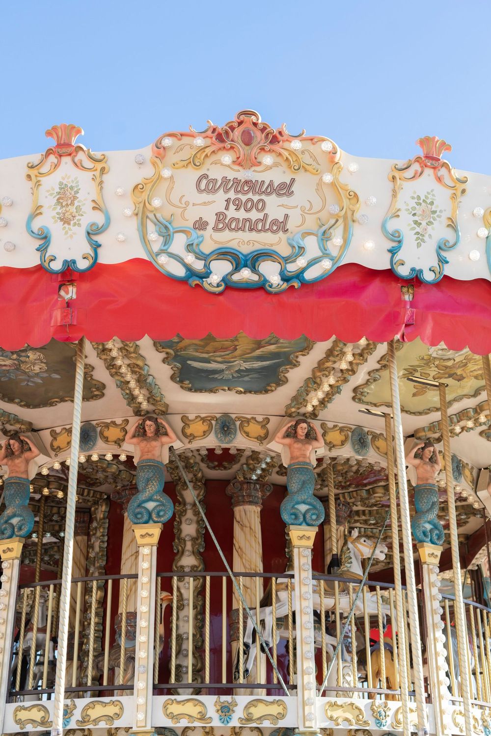 A vintage carousel with ornate turquoise and gold decorations featuring a red canopy and baroque-style design details against blue sky.