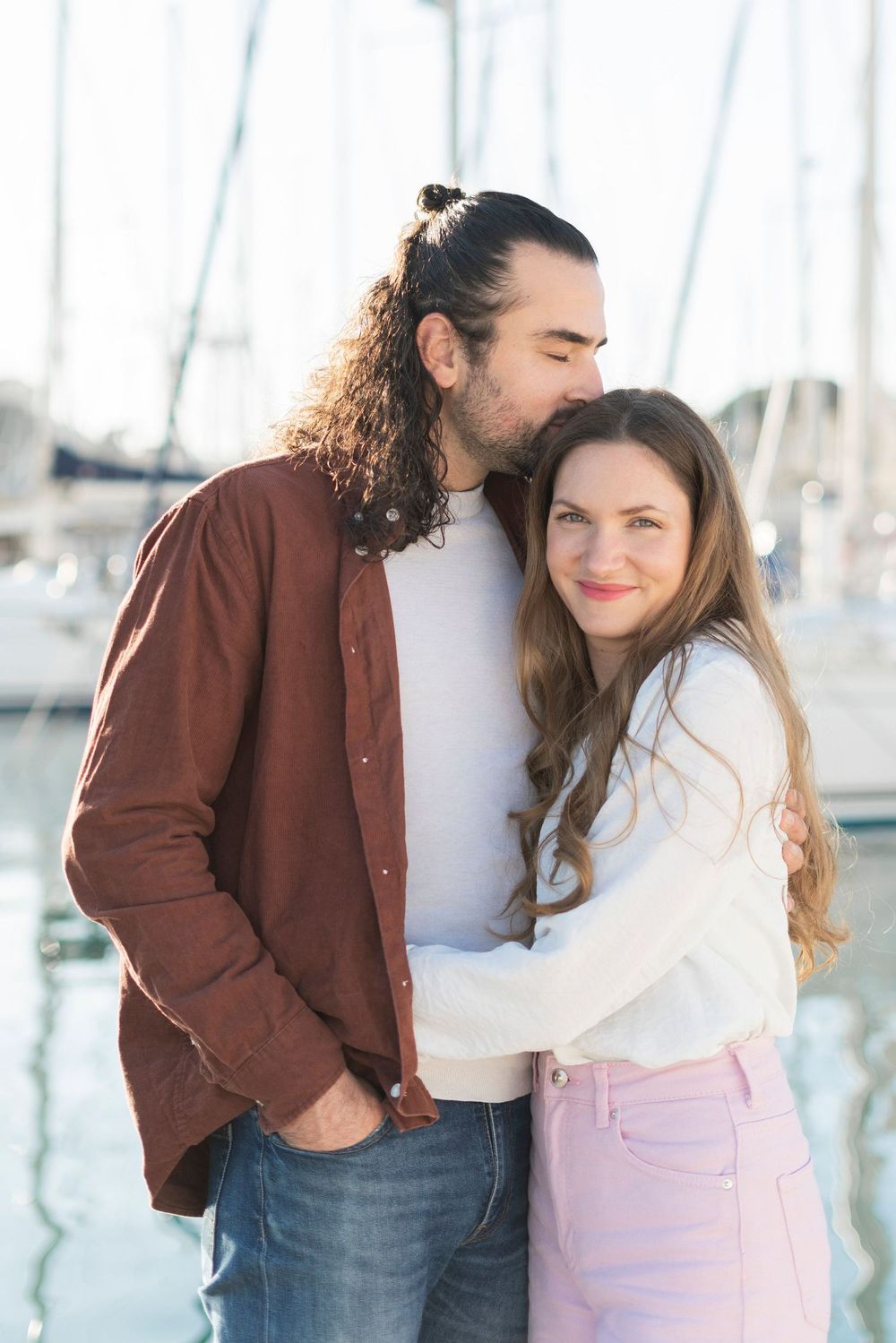 A couple embraces at a marina with sailboats in the background on a bright sunny day.
