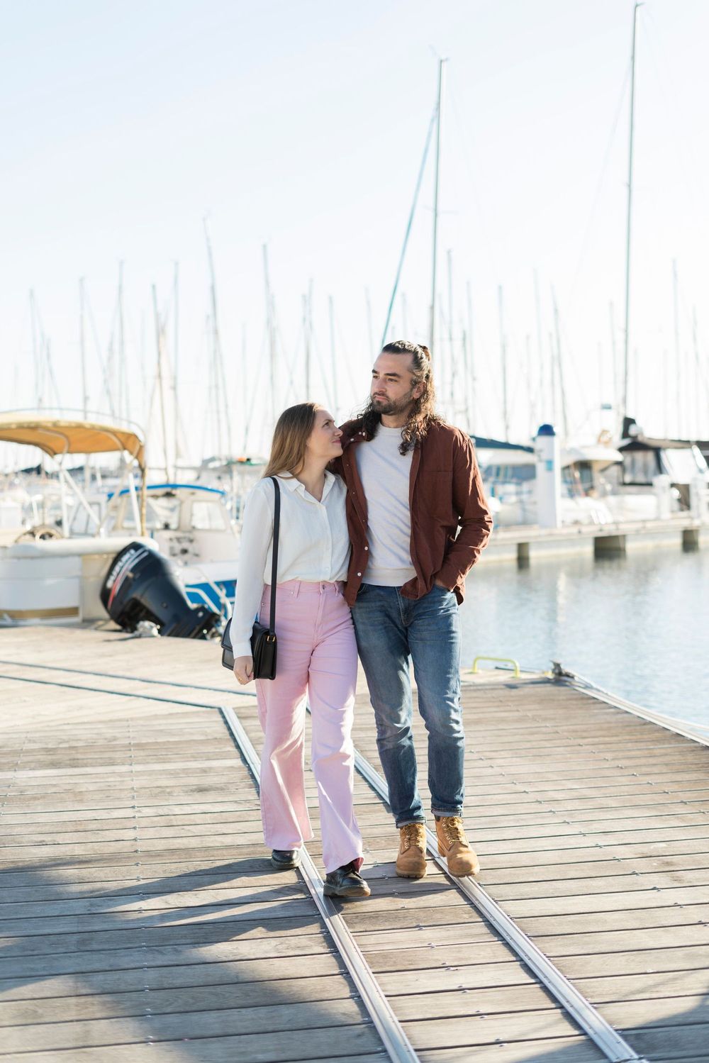 Couple standing on wooden marina dock surrounded by sailboat masts on a sunny day.