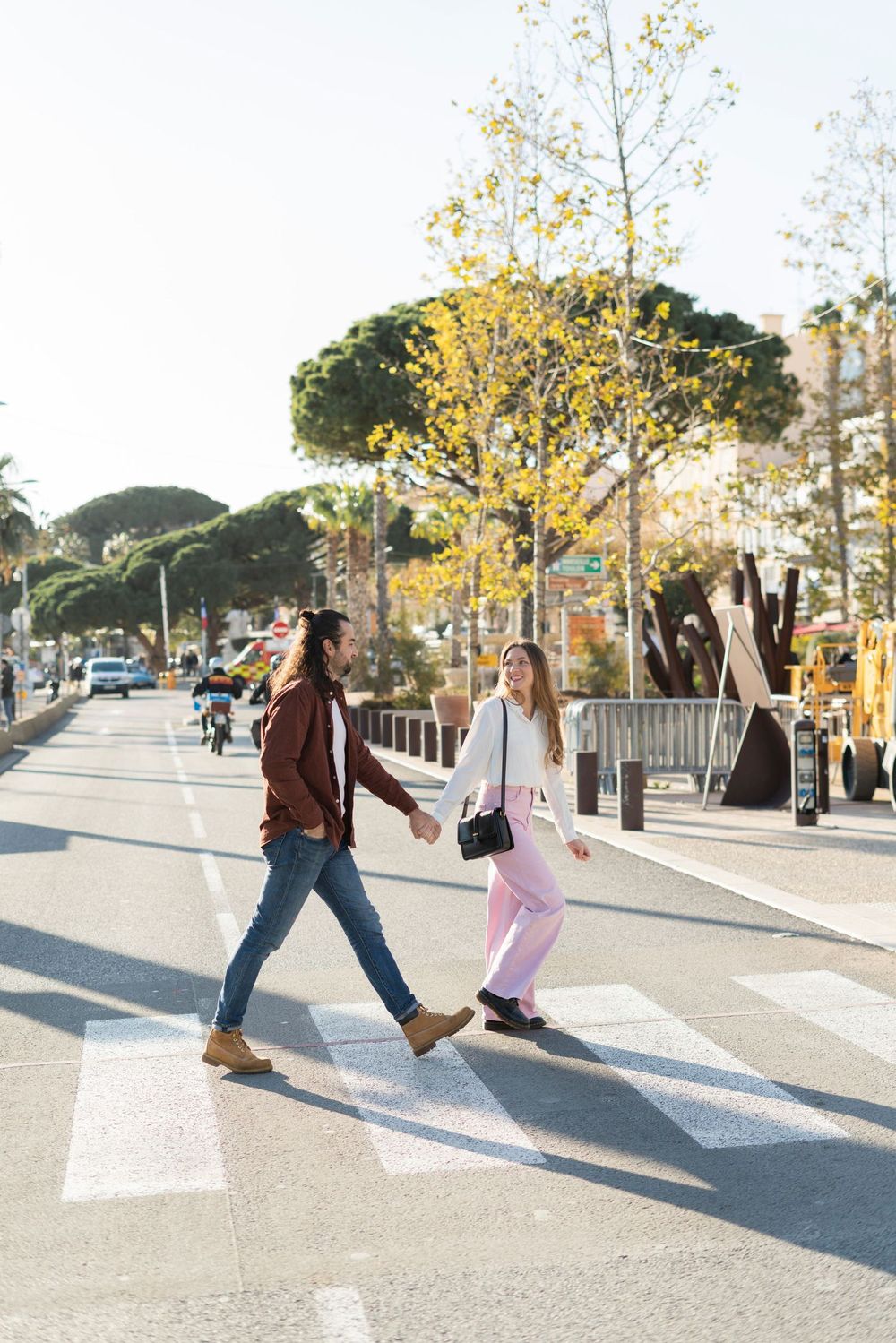 Two people holding hands while crossing a sunny street with trees and playground equipment in the background.