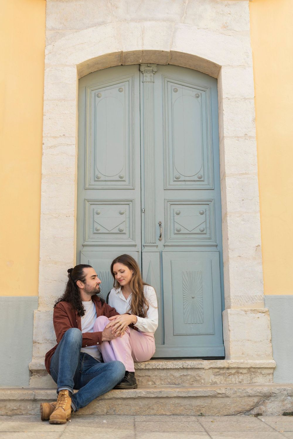 A couple sits together on stone steps in front of a light blue vintage door against a yellow building.