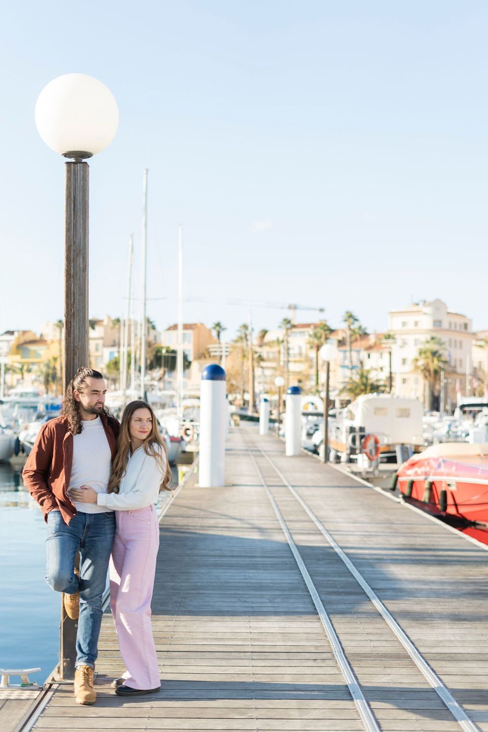 A couple stands together on a wooden pier with boats and palm trees in the marina background during sunset.