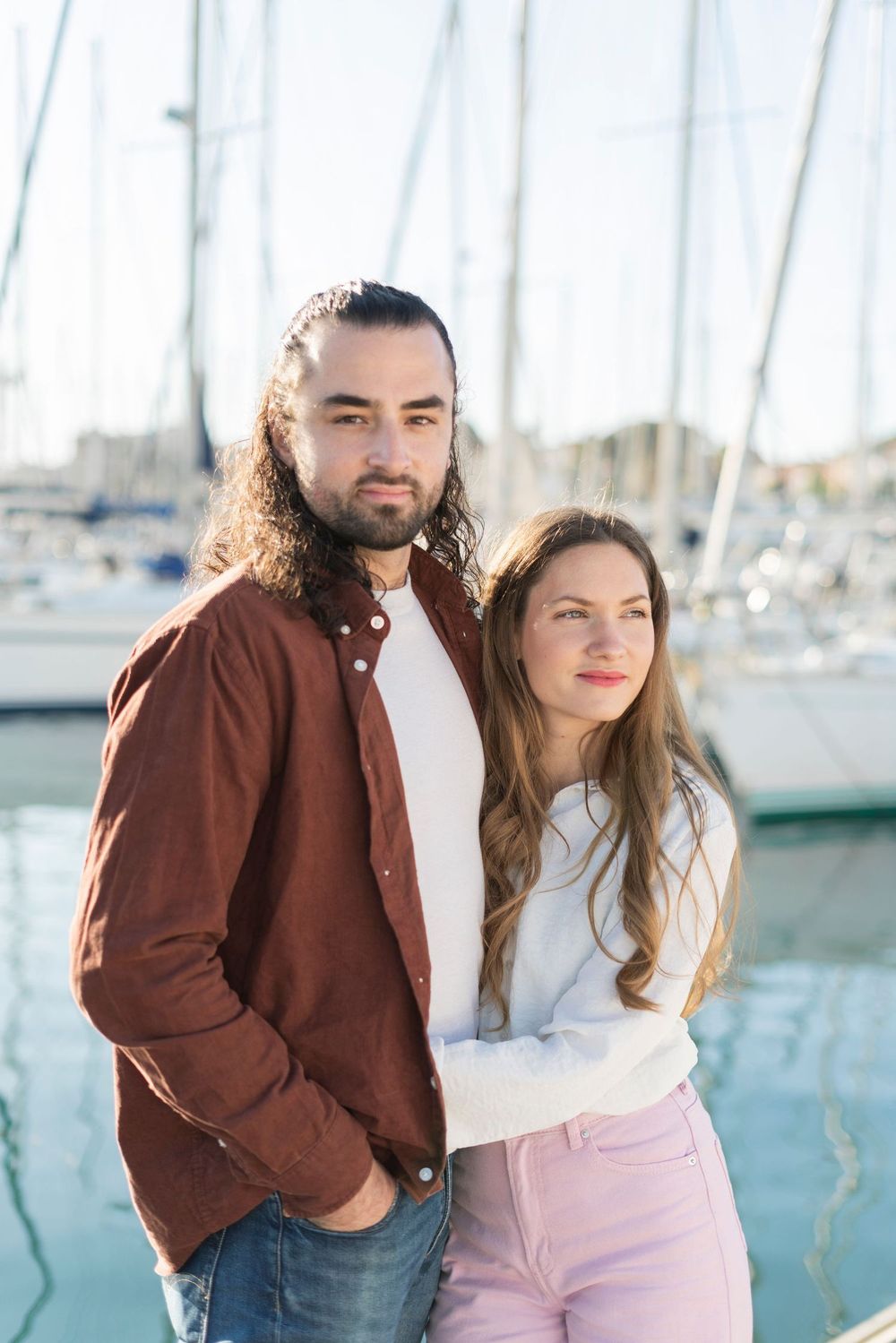 A couple stands together at a marina with boats in the background, wearing casual attire on a sunny day.
