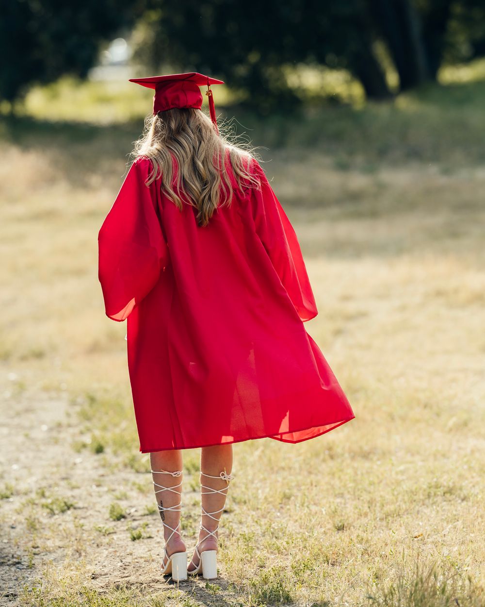 Graduate in flowing red cap and gown stands in a sunlit field wearing strappy sandals.