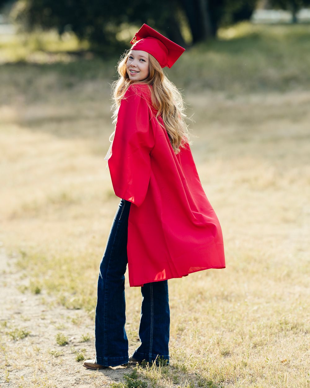Graduate wearing red academic regalia poses in casual jeans against a natural outdoor backdrop.