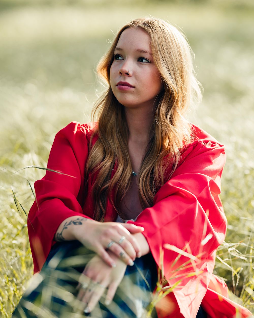 Graduate in bright red academic regalia sits thoughtfully in a sunlit meadow.