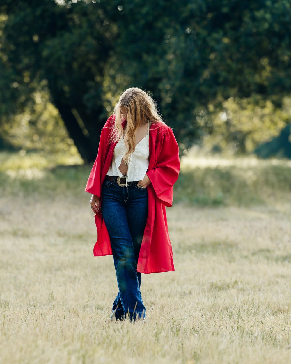Graduate in red gown and casual attire walks through a sunny field.