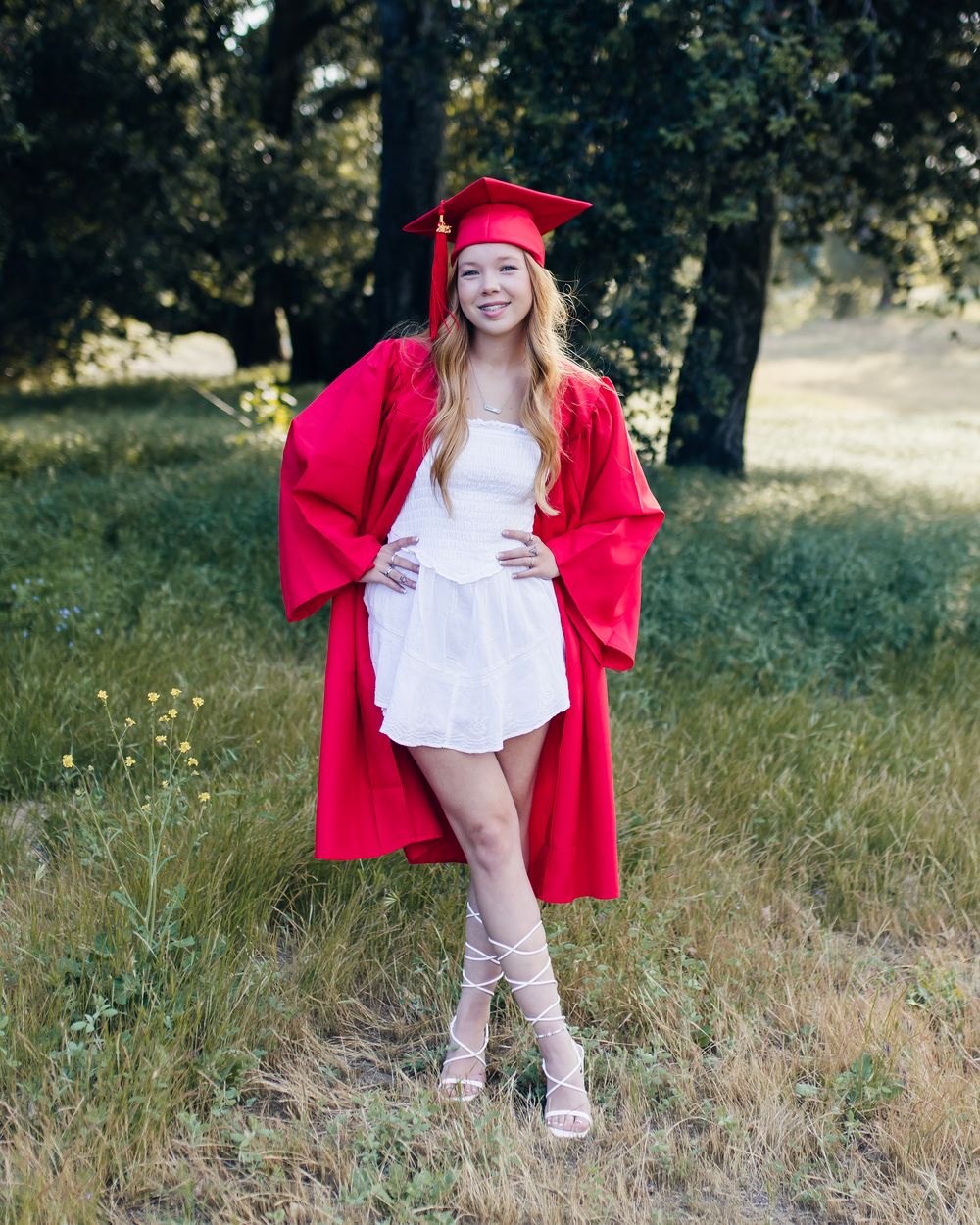 A figure in a bright red graduation gown poses playfully in a sunlit field.