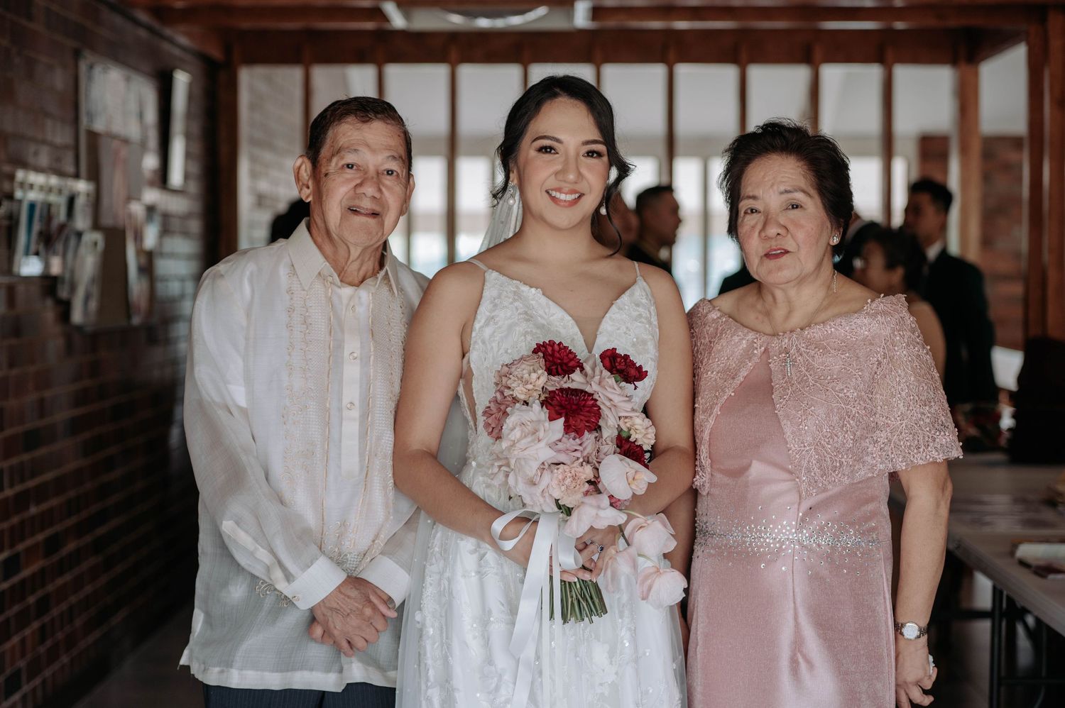Three people pose together at a wedding celebration with white and pink formal attire and a red and white bouquet.