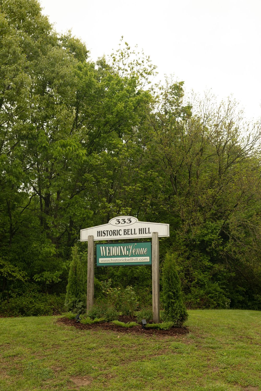 A wooden sign for Historic Bell Mill surrounded by lush green trees and landscaping.