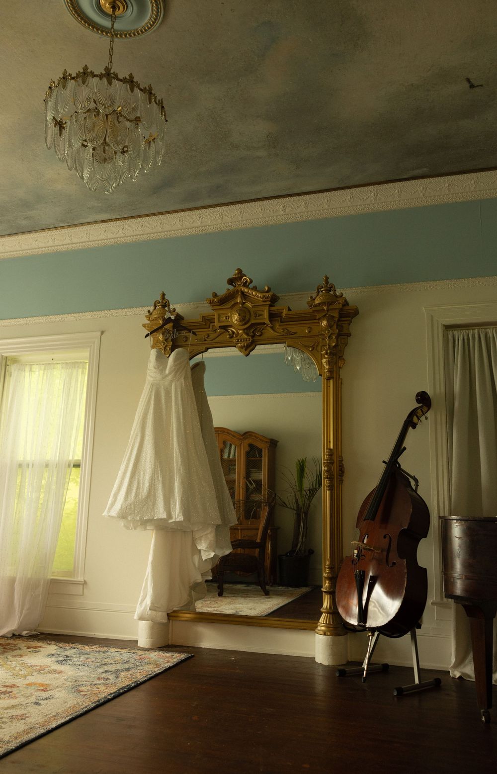 White wedding dress hanging on ornate gold mirror with cello in elegant room.