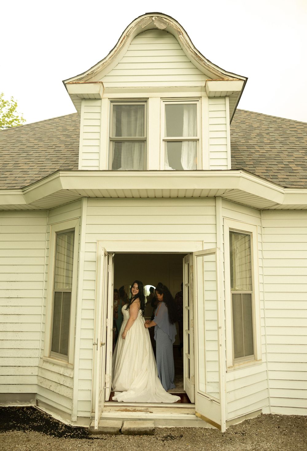A classic white farmhouse porch with curved dormer window and wedding party silhouetted in doorway entrance.