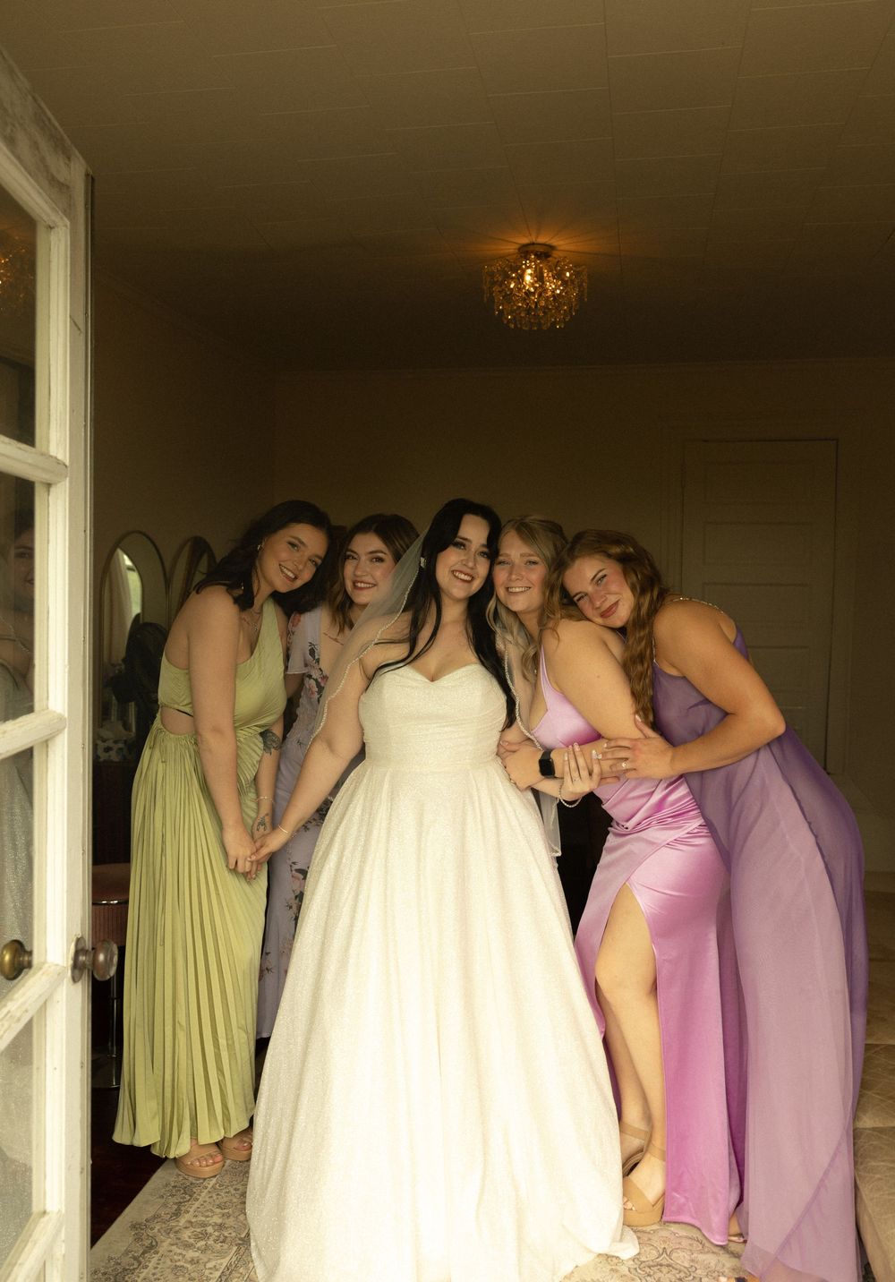 A bride in a white gown poses with bridesmaids wearing colorful dresses in varying shades of purple, pink and green.