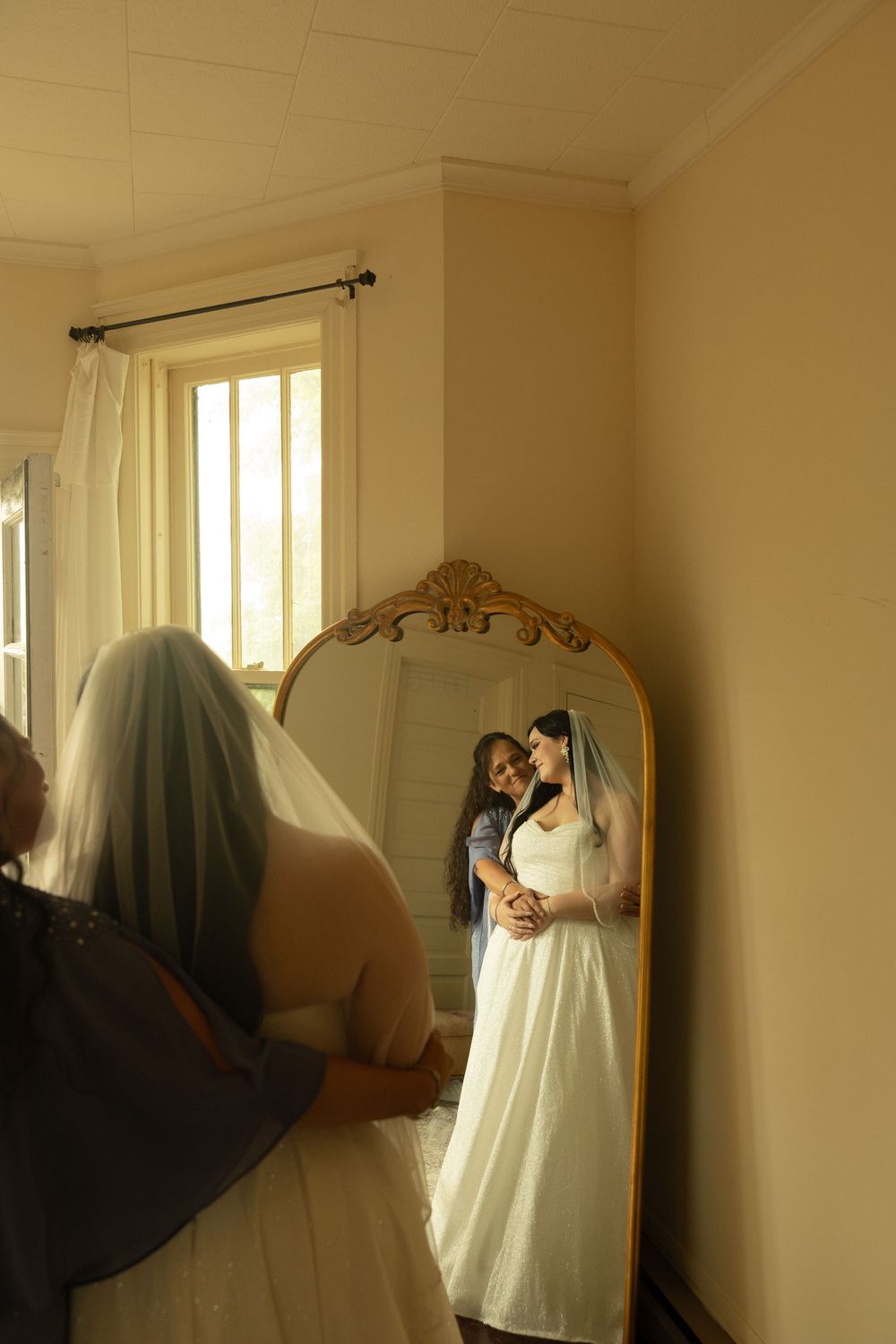 A wedding dress reflection captured in an ornate gold-framed mirror beside a window with natural light streaming in.