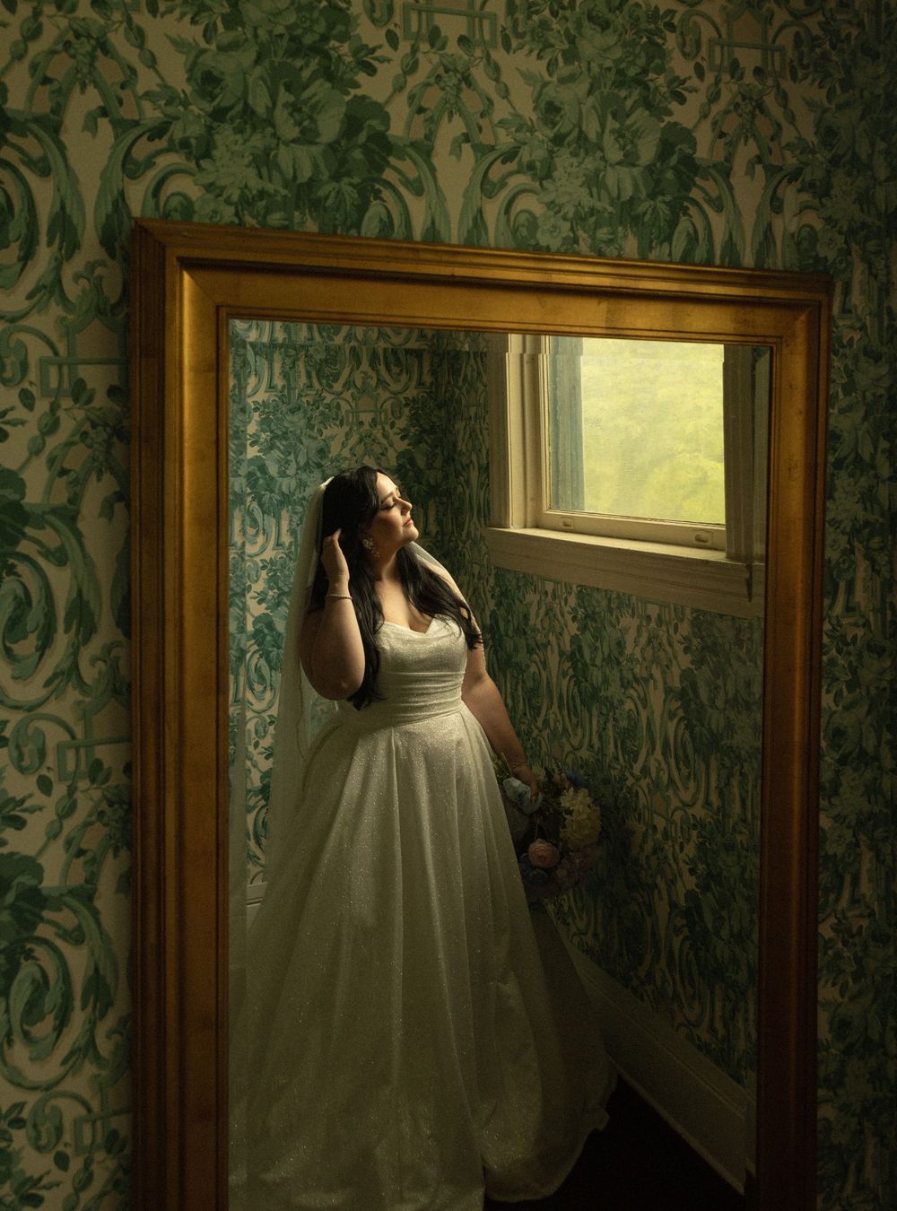 A silhouetted figure in a white wedding dress poses by a window against ornate vintage wallpaper.