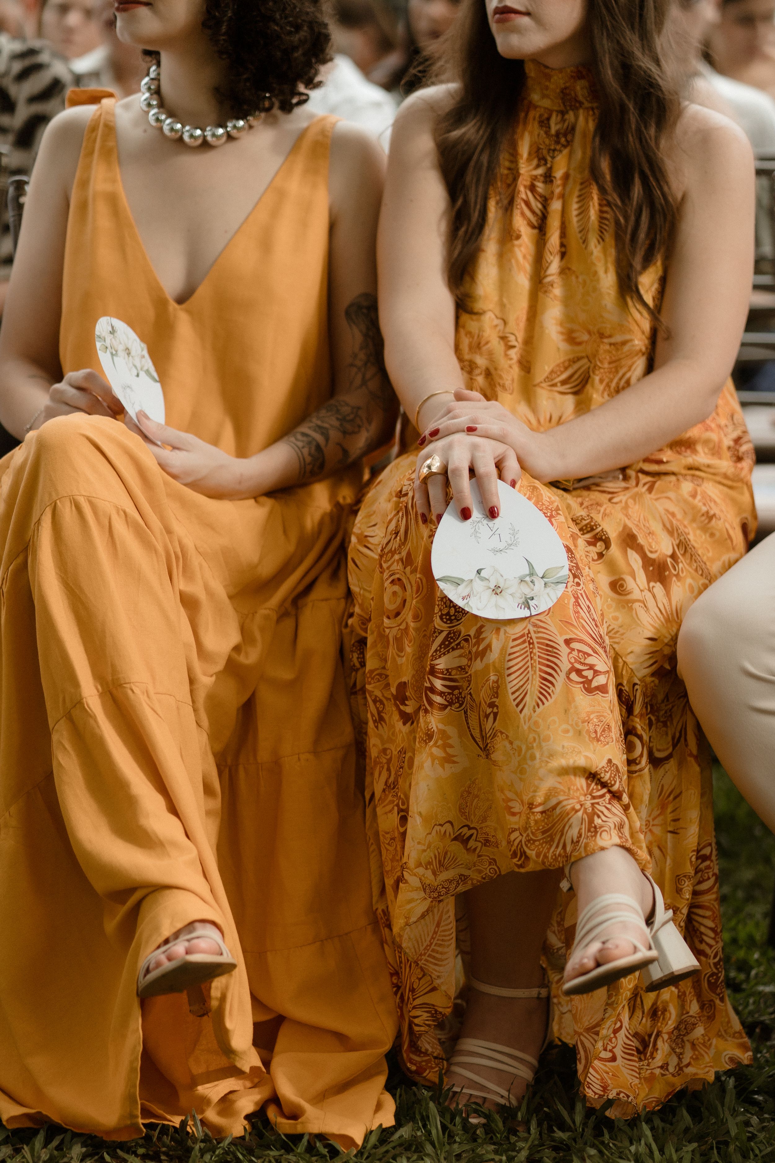 Wedding guests in flowing orange and yellow dresses holding decorative fans while seated.