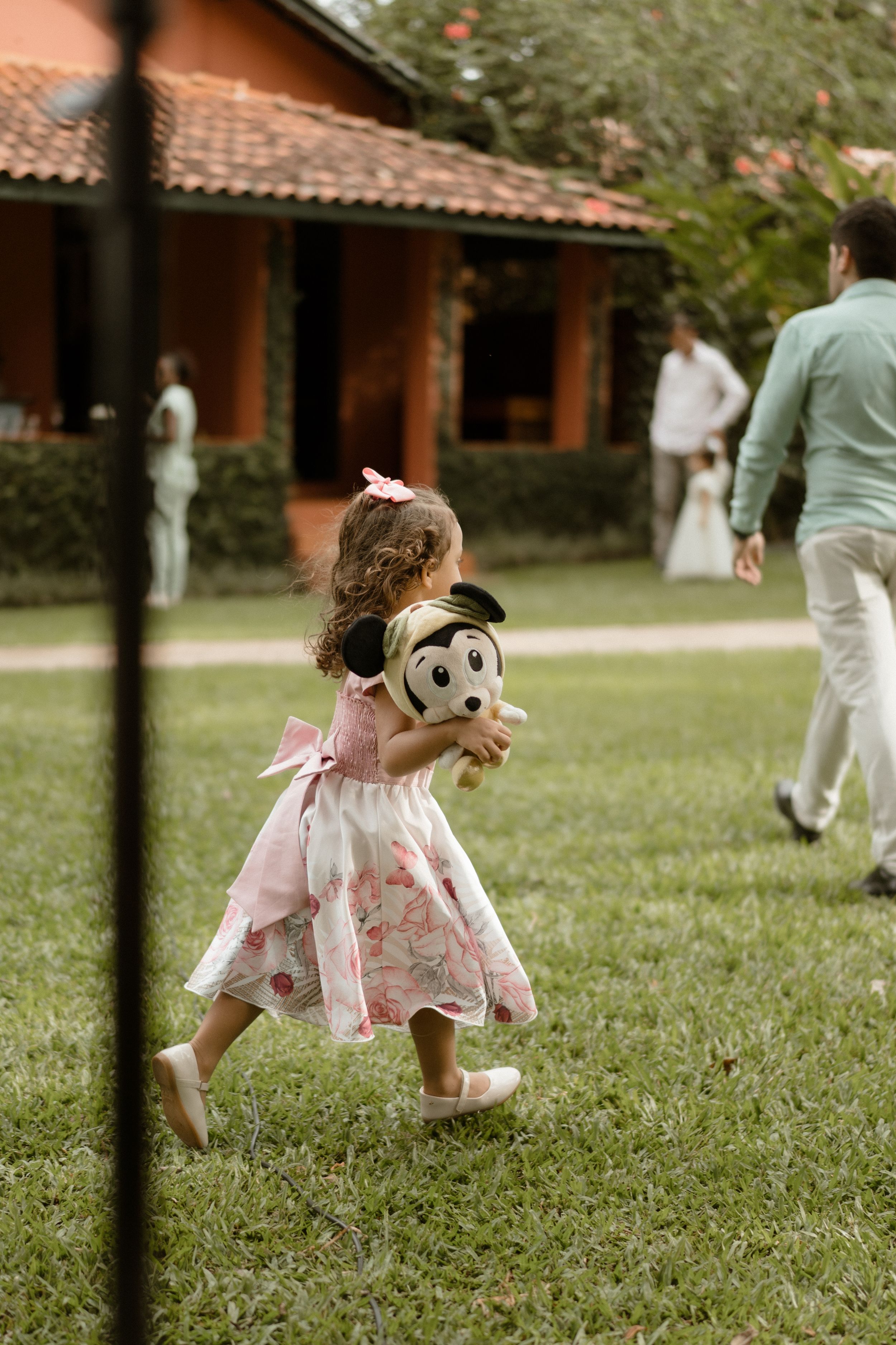 A small child in a pink dress runs across a lawn carrying a stuffed toy during a wedding.