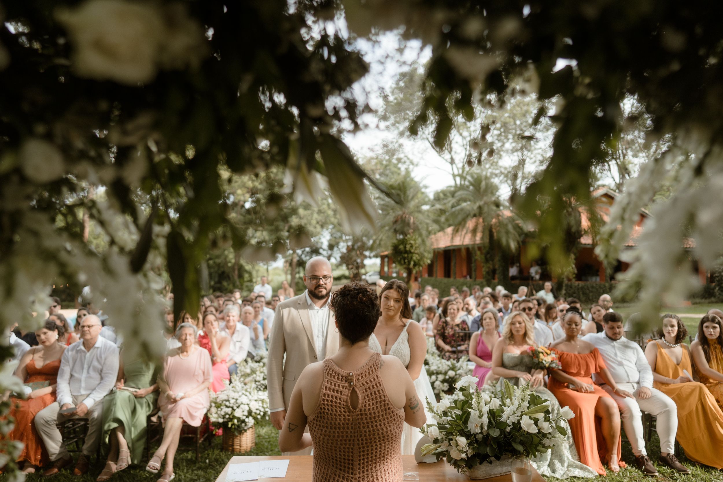 A garden wedding ceremony with guests seated on white chairs surrounded by lush greenery.