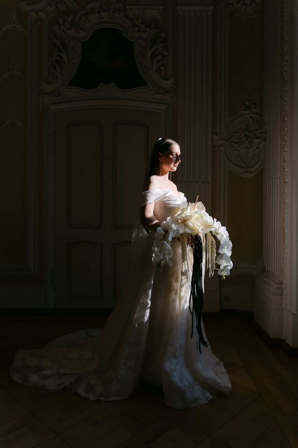 A bride in an elegant white off-shoulder gown holding a luxurious bouquet of white flowers stands in a grand ornate room.