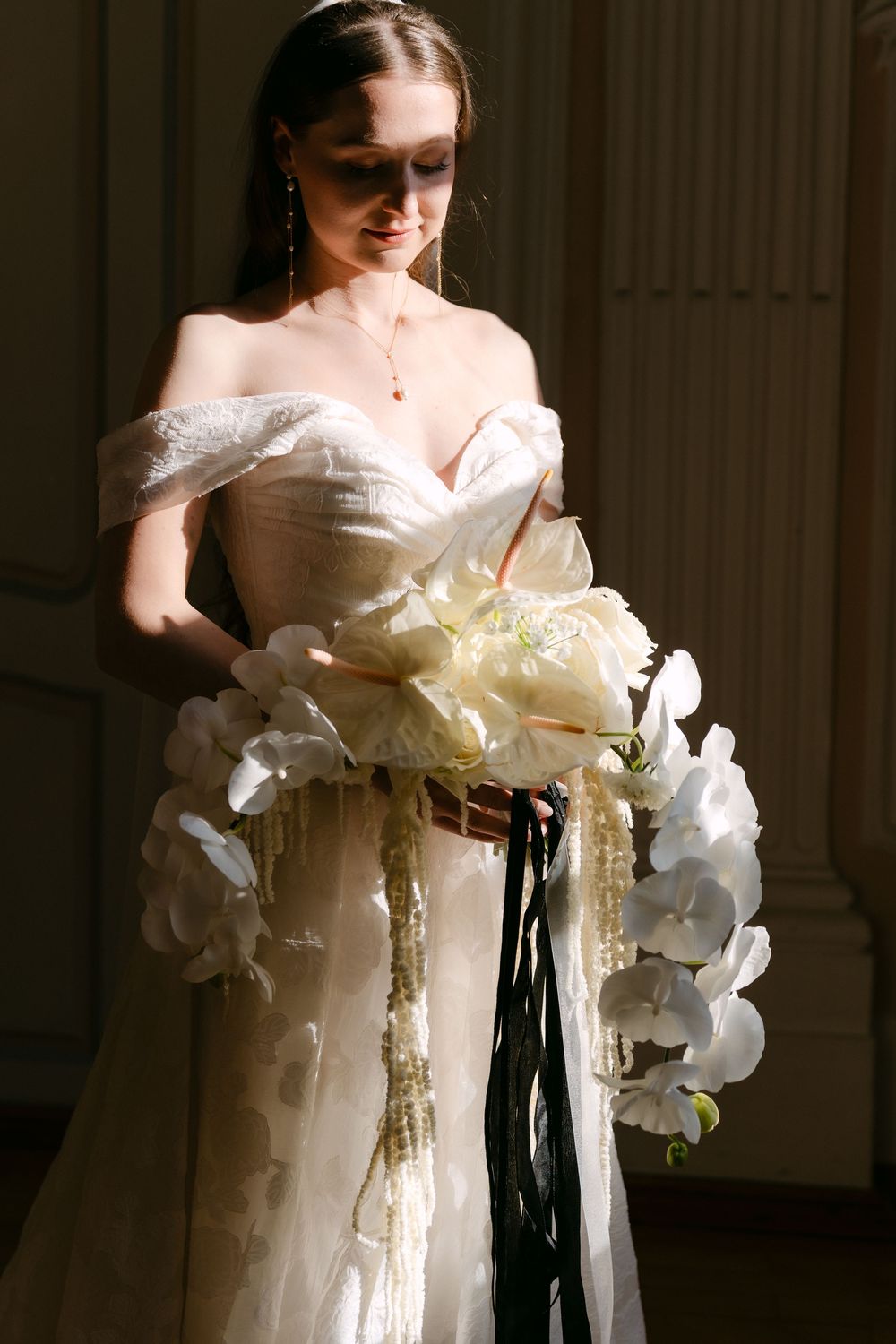 Bride in an elegant off-shoulder white wedding gown holds a cascading bouquet of white orchids with black ribbon accents.