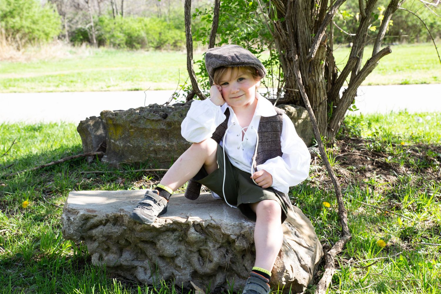 Child in vintage costume rests on stone in sunny garden setting.