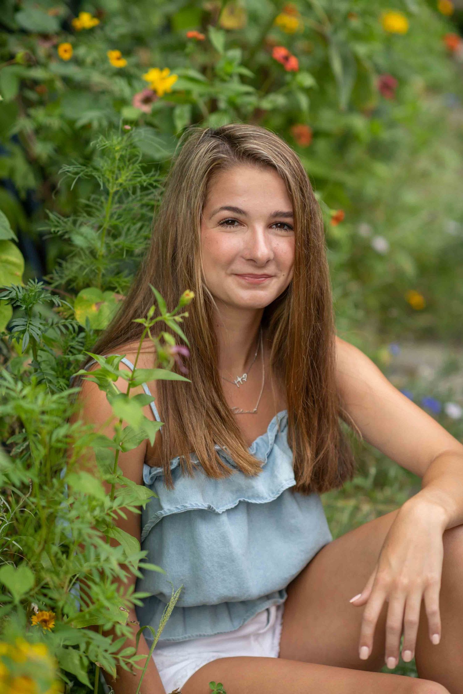 Young woman with long brown hair smiling in a garden surrounded by colorful flowers, wearing a blue ruffled top.