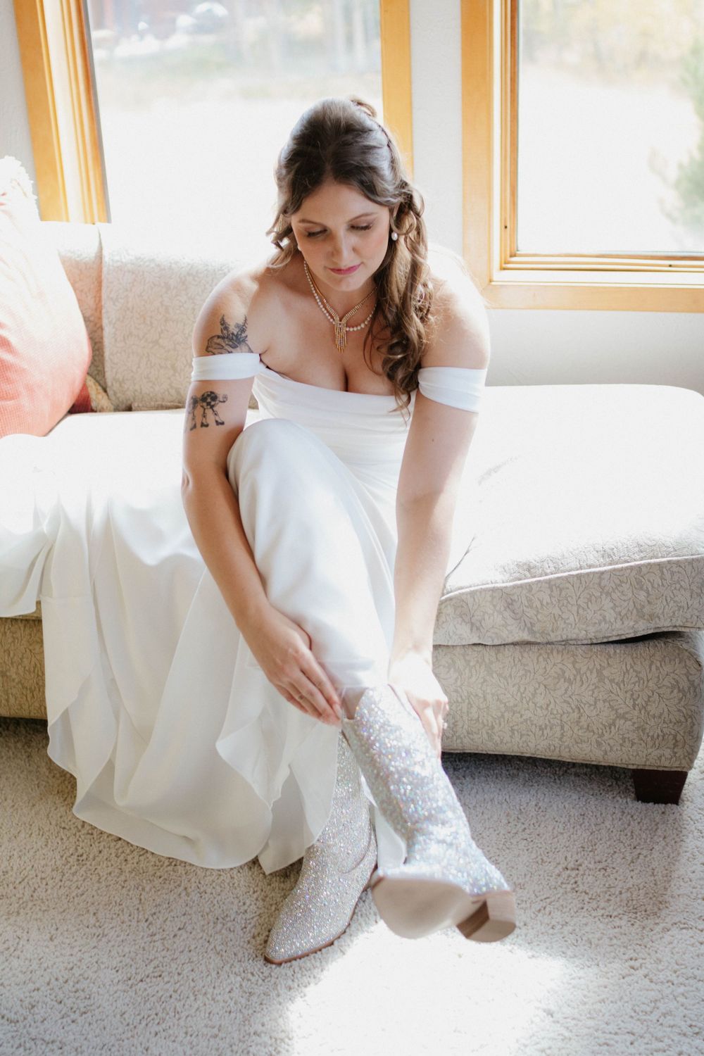 A bride in a white wedding dress adjusts her lace garter while sitting on a light colored sofa in a sunlit room.