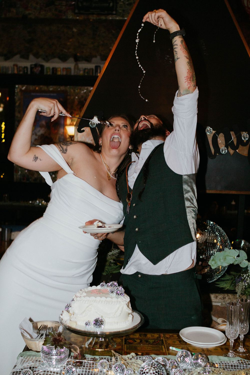Newlyweds celebrate enthusiastically at their wedding reception table with a white cake and decorative table settings.