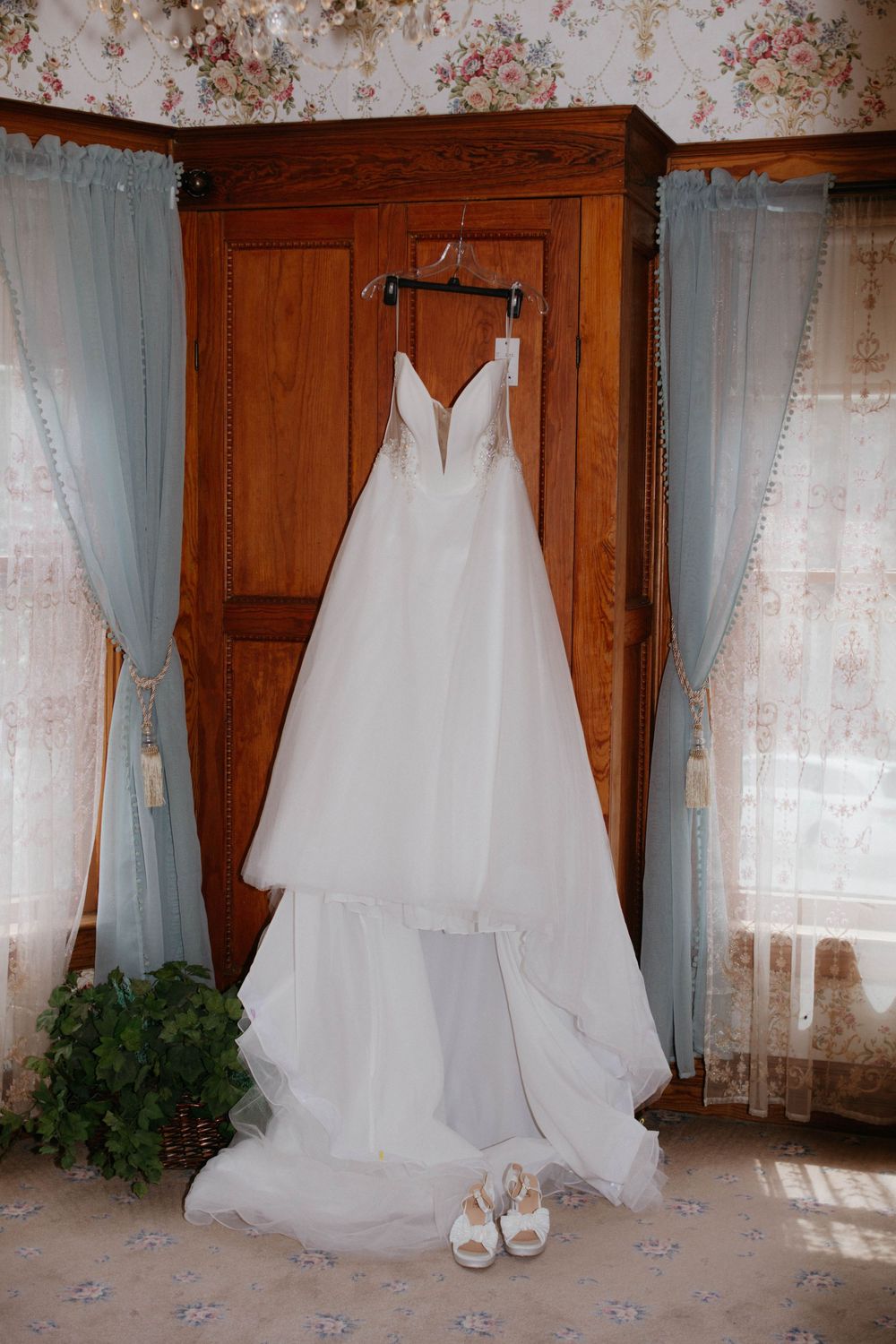 A white wedding dress with plunging neckline hangs on wooden wardrobe with matching shoes beneath and sheer curtains.