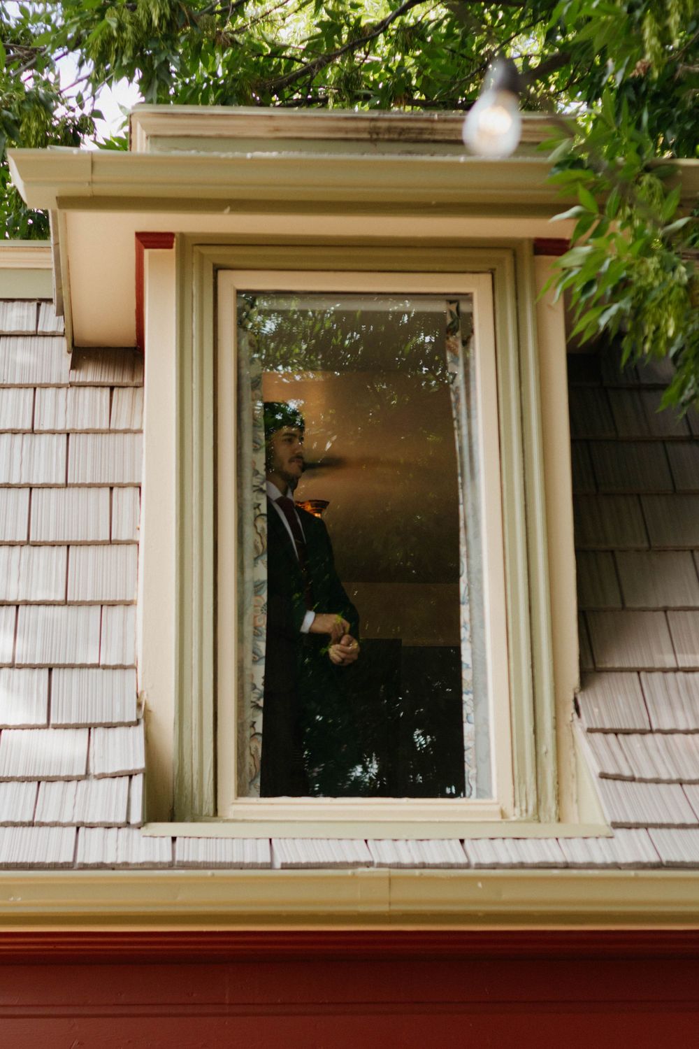A cat sits in the window of a house with white shingle siding and cream trim, peering outside through the glass.