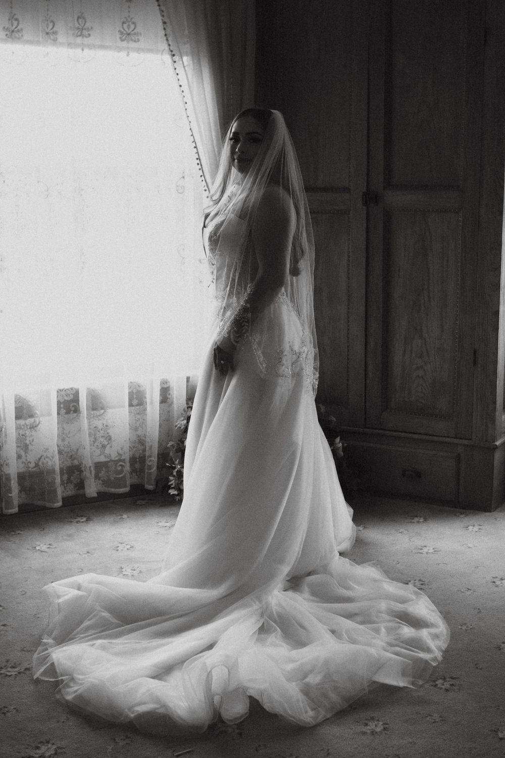 A dramatic black and white photo of a bride in a flowing wedding gown with veil standing by a window in vintage style.