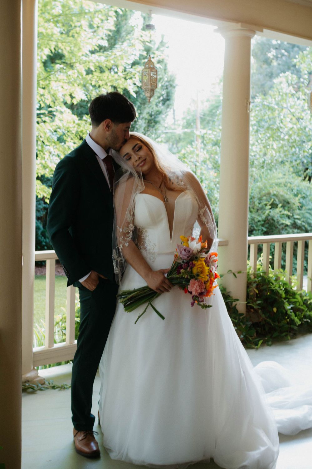 Romantic wedding moment captured on an elegant white columned porch surrounded by lush greenery.