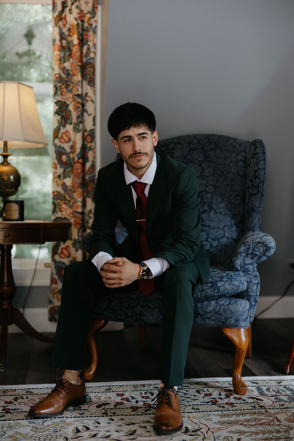 Person in dark suit sits on blue velvet couch while looking at mobile phone in moody interior setting.