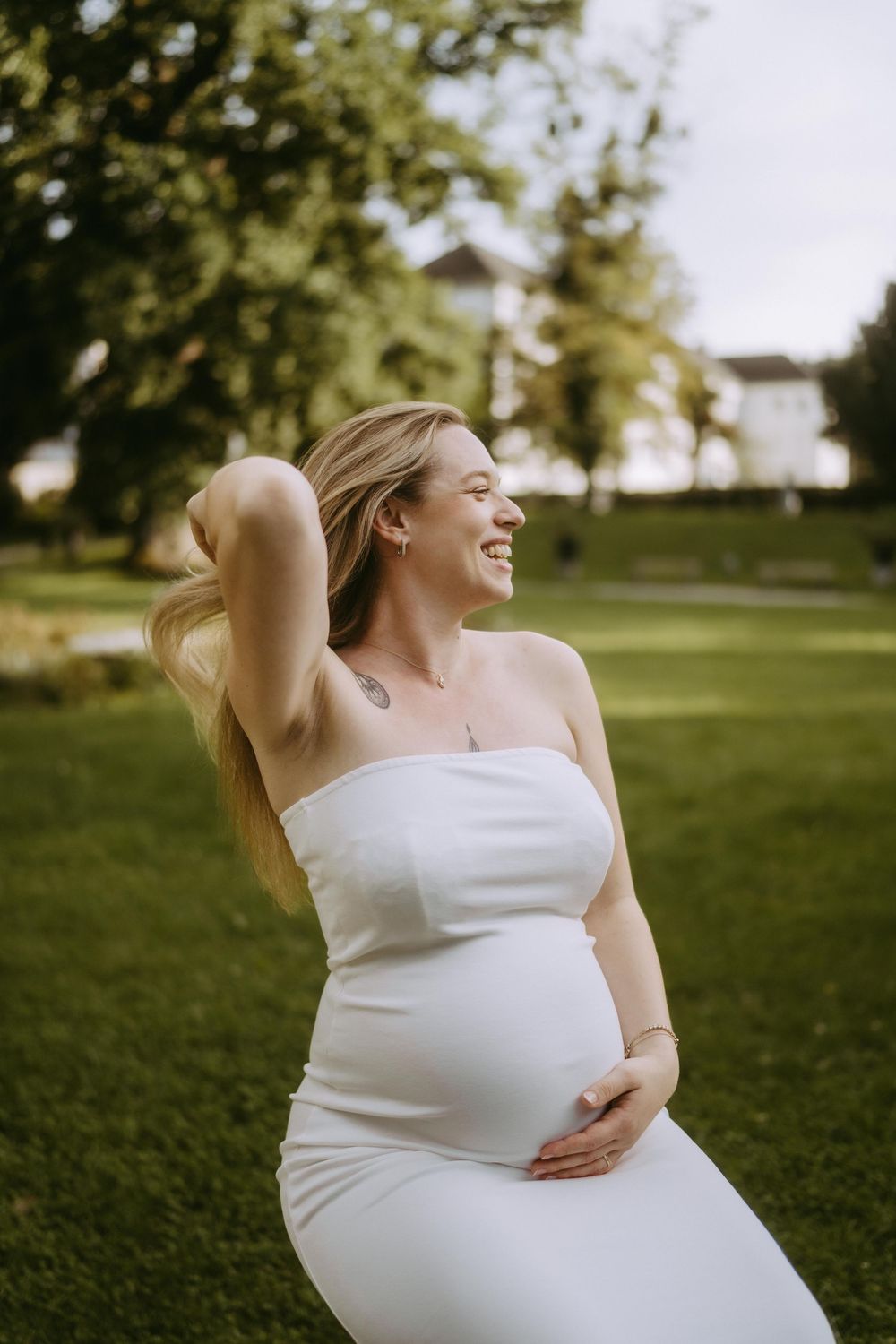 A woman in a white dress cradles her baby bump in a sunny outdoor setting.