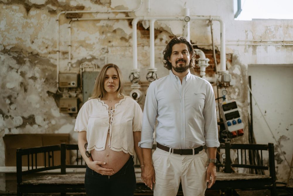 A romantic couple poses against a rustic wall in casual white outfits during a photo session.
