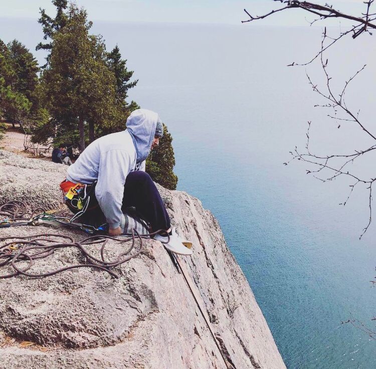 Person in gray hoodie sitting precariously on edge of steep cliff overlooking blue waters below.