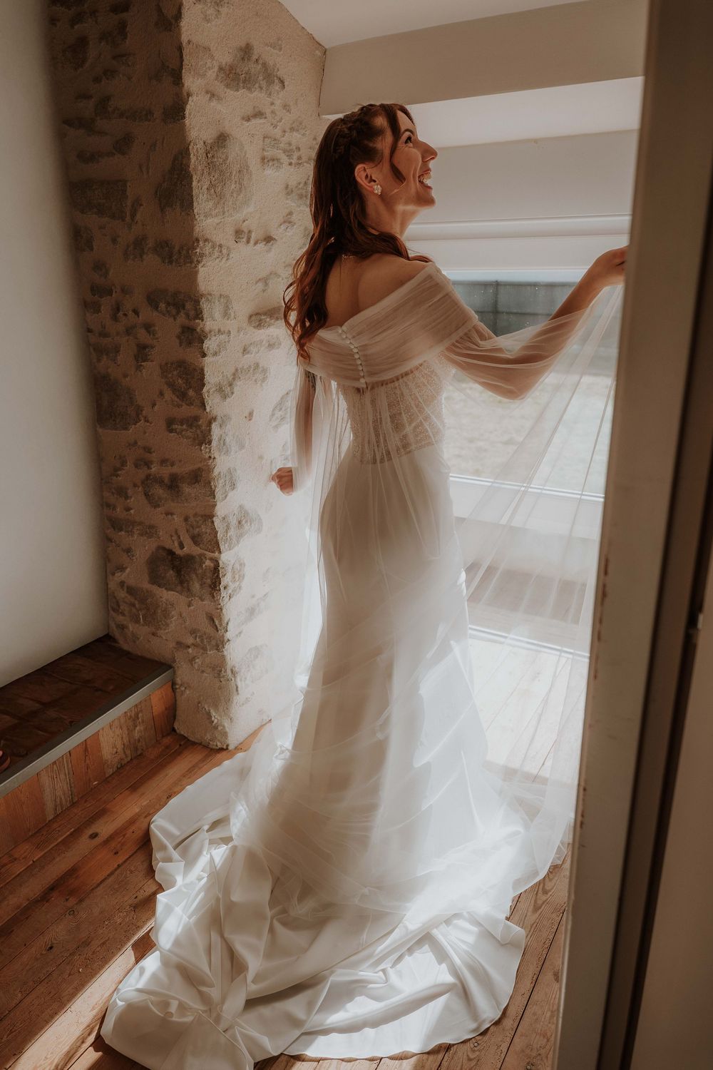 Bride in off-shoulder white wedding gown with flowing train stands by window in rustic stone room.