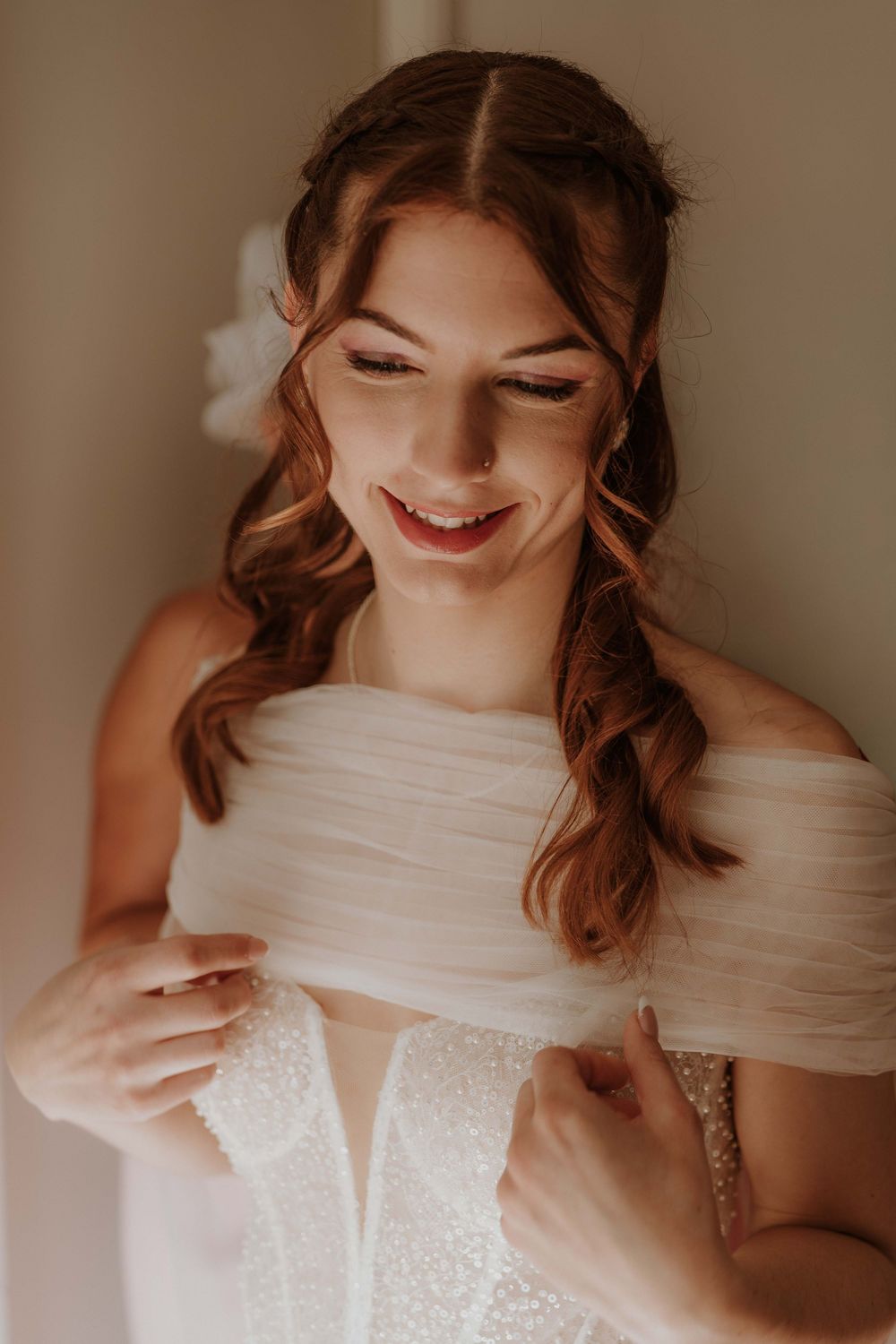 Smiling bride in a white off-shoulder wedding dress with floral hair accessory during bridal preparation.