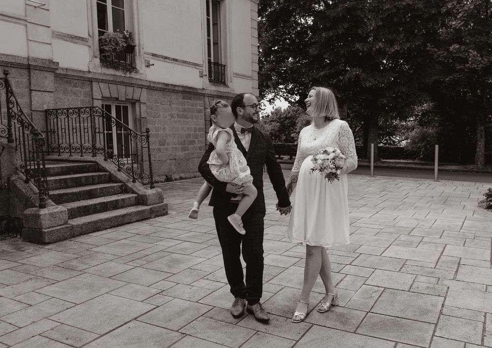 Black and white photo of a couple in wedding attire holding hands, man carrying a child, walking outside a stone building.
