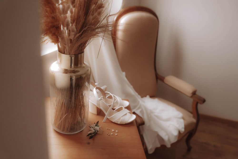 A delicate white wedding dress draped over a vintage chair next to a vase of dried pampas grass in warm lighting.