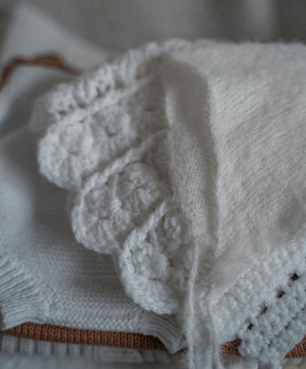 Soft white baby bonnet resting on folded knitted blankets, styled for a newborn lifestyle photography session in Derbyshire.