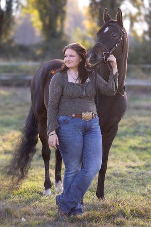 A person in jeans and gray sweater stands beside a dark brown horse in a grassy field at sunset.
