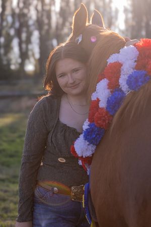 Horse wearing red, white and blue patriotic flowers stands with rider in sunlit field at golden hour.