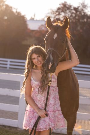 A woman in a floral pink dress poses with a dark brown horse at sunset by a white fence.