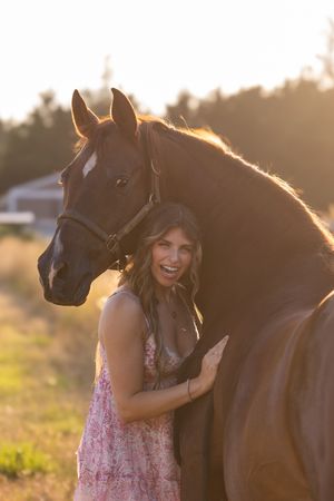 A woman in a pink floral dress embraces a dark brown horse during a golden sunset photoshoot.