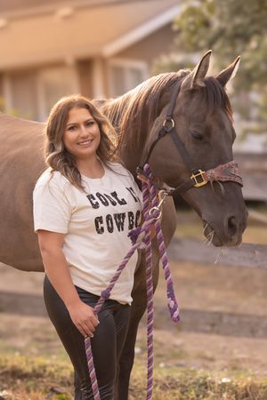 A person in a white t-shirt stands next to a dark brown horse with a purple lead rope at sunset.