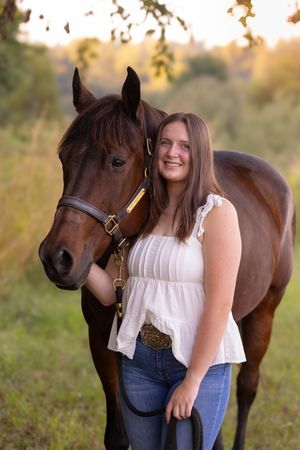 A beautiful brown horse stands with someone in a white top and jeans during a golden sunset photo session.