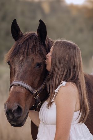 A woman in a white dress shares a tender moment with a brown horse wearing a bridle in a sunlit field.