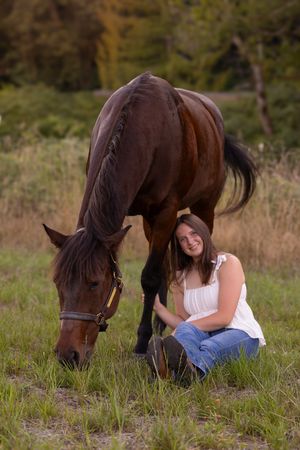 A dark brown horse grazes in a field while someone sits nearby in casual attire during a peaceful outdoor moment.